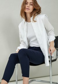 Bílá košile s knoflíky, oblečená přes bílý top bez rukávů, v kombinaci s námořnickými, úzkými kalhotami. Modelka sedí na moderní černé židli.