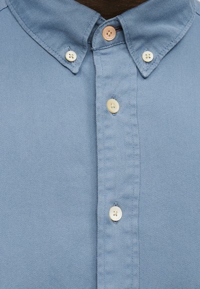 Ανοιχτό μπλε πουκάμισο με κλασικό γιακά, ελαφρώς υφασμένο ύφασμα και μπεζ και λευκά κουμπιά. Λεπτομέρεια της περιοχής του λαιμού εμφανής.