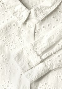 Valkoinen broderad paita, jossa on kaulus, napitus ja herkät kukka- ja pyöreät kuviot kankaassa. Pehmeä rakenne.