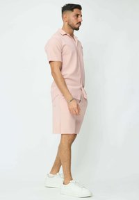 Chemise à manches courtes rose clair et shorts assortis, texture côtelée, coupe décontractée, associée à des baskets blanches, posant sur fond uni.