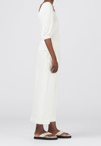 Weiße, texturierte lange Rock kombiniert mit einer taillierten weißen Bluse mit Puffärmeln. Das Model trägt einfache flache Sandalen mit kontrastierenden Riemen.