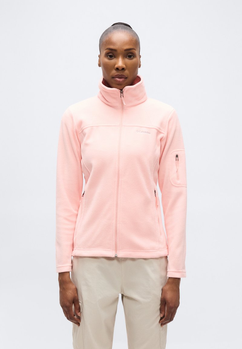 Γυναίκα που φοράει γούνινο μπουφάν με φερμουάρ σε ανοιχτό ροζ χρώμα και ψηλό γιακά, με μπεζ παντελόνι, στέκεται μπροστά σε απλό λευκό φόντο.