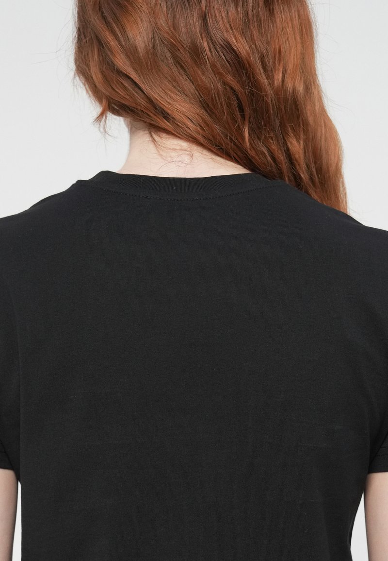 Persona con lunghi capelli rossi che indossa una maglietta nera semplice, vista di spalle su uno sfondo chiaro e uniforme.