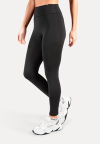 Leggings nere a vita alta realizzate in materiale liscio ed elastico. Indossate con scarpe da ginnastica bianche con dettagli neri.