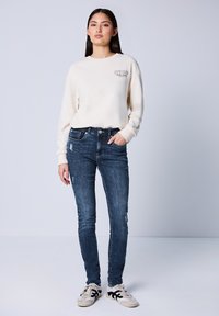 Kremi t-paita logolla, yhdistetty tummansinihin slim fit -farkkuihin, joissa on kuluneita kohtia. Mustilla yksityiskohdilla varustetut sneakerit täydentävät asun.