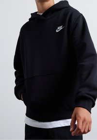 Černá mikina s kapucí z jemné tkaniny, s klokaní kapsou, žebrovanými manžetami a bílým logem Nike na hrudi. Přes bílou košili.