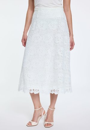 Weißer Spitzenrock mittlerer Länge mit Blumenmuster, getragen mit weißem Oberteil und beigen Sandaletten mit hohen Absätzen und Knöchelriemen.