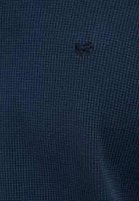 Niebieska fakturowana tkanina z wzorem wafla oraz małym czarnym logo zwierzęcia wyszytym po lewej stronie.