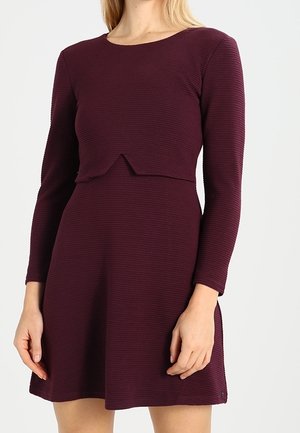 Kobieta w ciemnofioletowej, prążkowanej sukience z długim rękawem, dopasowaną w talii, z małym przodem zachodzącym na siebie.