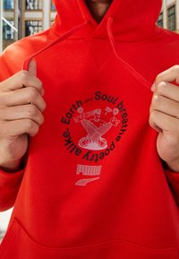 Röd hoodie i bomullsblandning, med en grafik av en skateboardåkare med blommor och text. Utrustad med kenguruficka och dragsko i huvan.