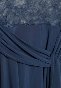 Sötétkék ruha virágmintás csipkebordűrrel és sima anyagú szoknyával, széles övvel, amely csomóra van kötve a deréknál.