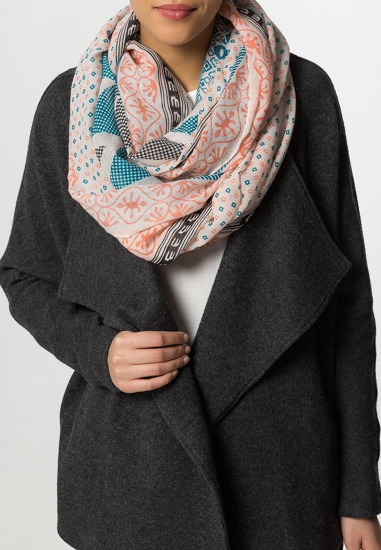 Femme portant un manteau gris foncé et une grande écharpe à motifs avec des motifs géométriques et floraux pêche, bleus et noirs enroulée autour de son cou.