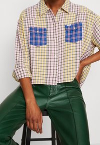 Karierte Hemdbluse in Pink, Gelb und Grün mit zwei blauen karierten Brusttaschen; kombiniert mit grünen Lederhosen.