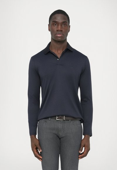 Polo bleu marine à manches longues avec col, doté d'une patte de boutonnage et d'un petit logo sur la poitrine, associé à un jean gris coupe slim.