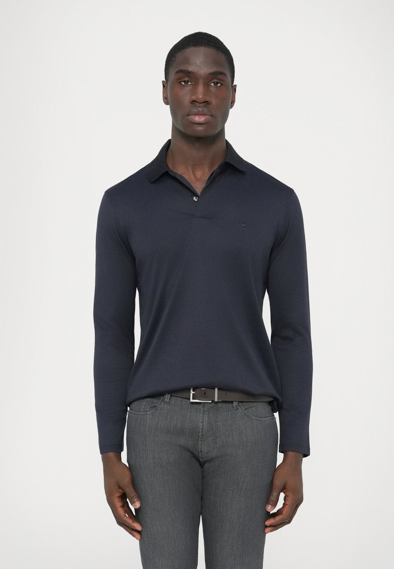 Tengerészkék hosszú ujjú galléros póló, gombos sliccel és kis logóval a mellkason, szürke, karcsúsított szabású farmerral párosítva.