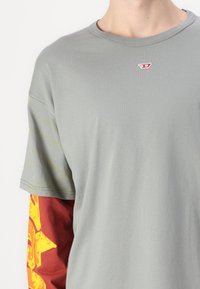 T-shirt grigio a maniche corte con collo rotondo, caratterizzato da un logo rosso sul petto e una lunga manica rossa con un dettaglio grafico floreale giallo.