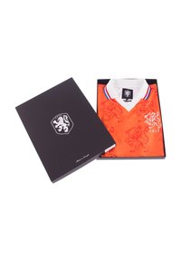 Oranje voetbalshirt met leeuwenpatroon, witte kraag en blauw-rood accent. Verpakt in een zwarte doos met een leeuwembleem en handtekening.