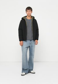 Black puffer jacket with a green lining, grey sweater, and flared light blue jeans. The model stands against a neutral background.