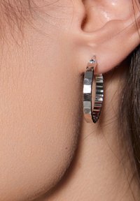 Srebrna kolczyka na kółku z teksturowanym, żłobkowanym wzorem i błyszczącym wykończeniem. Kolczyka zawiera mały, przezroczysty akcent w postaci kamienia szlachetnego.