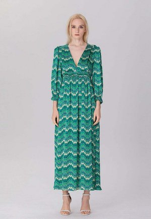 Femme portant une robe longue à manches longues verte à motif zigzag, avec un profond décolleté en V et des sandales blanches à talons hauts, debout sur un fond uni.