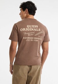 Bruine katoenen T-shirt met een crèmekleurige graphic op de achterkant, inclusief "GUESS ORIGINALS", een schroefillustratie en contactinformatie.