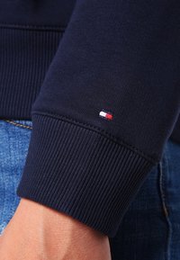 Polsino di felpa blu navy con texture a coste, caratterizzato da un piccolo logo tricolore (rosso, bianco, blu) ricamato sull'orlo.