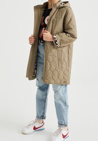 Chaqueta acolchada de color verde oliva con forro de leopardo, capucha y cremallera, combinada con jeans desgastados de color azul claro y zapatillas blancas con rojo.