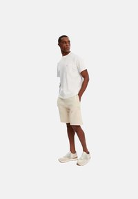 Witte katoenen t-shirt met korte mouwen, beige shorts en lichtgrijze sneakers met designelementen. Staande pose met handen in de zakken.