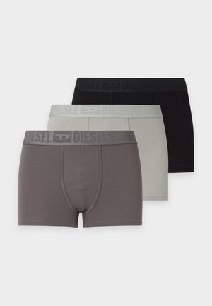 Jeune homme aux cheveux courts et foncés, debout pieds nus, portant un boxer gris clair sur un fond blanc uni.