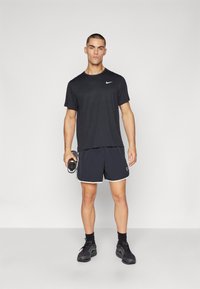 Svart Nike T-skjorte og mørke shorts med hvite detaljer. Modellen holder en vannflaske og har på seg svarte joggesko. Minimalistisk design.