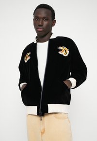 Um casaco bomber de veludo preto com cabeças de tigre bordadas, fecho com zíper e punhos e barra em creme canelado. Usado por cima de uma camisa branca.