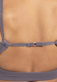 Grijze sportbeha van stof met een brede band en kruissende banden. Voorzien van metalen hardware en structuurelementen. Achteraanzicht toont de configuratie van de banden.