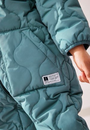 Kurtka z pikowanym wzorem w kolorze teal z gładką fakturą, widocznymi przeszyciami oraz metką naszytą po lewej stronie z instrukcjami pielęgnacji.