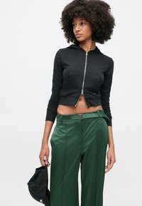 Μαύρο cropped φούτερ με φερμουάρ και κουκούλα, με μπροστινή τσέπη. Συνδυασμένο με πράσινο, πλισέ, ψηλόμεσο παντελόνι και μαύρη τσάντα.