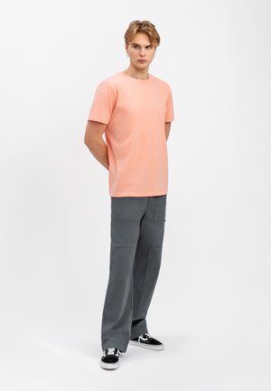 T-shirt di cotone color corallo chiaro con maniche corte abbinato a pantaloni grigi larghi con cuciture visibili e tasche, completato da sneakers nere.