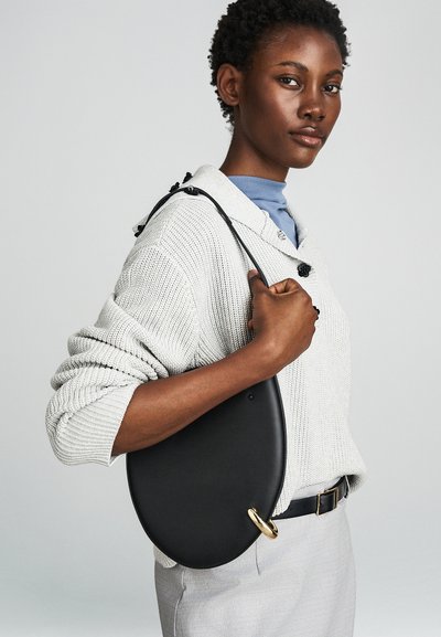 Fekete bőrtáska ovális formával, arany gyűrűs díszítéssel és levehető pánttal. Szürke pulóveren és kék ingben viselve.