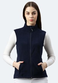 Marineblaue Fleece-Weste mit hohem Kragen, Reißverschluss und seitlichen Taschen, über einem weißen gerippten Langarmshirt getragen. Glatte Textur.