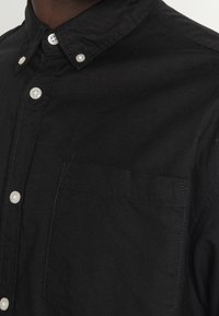 Camicia nera a bottone realizzata in tessuto testurizzato, dotata di una tasca sul petto, bottoni bianchi e un design di colletto classico.
