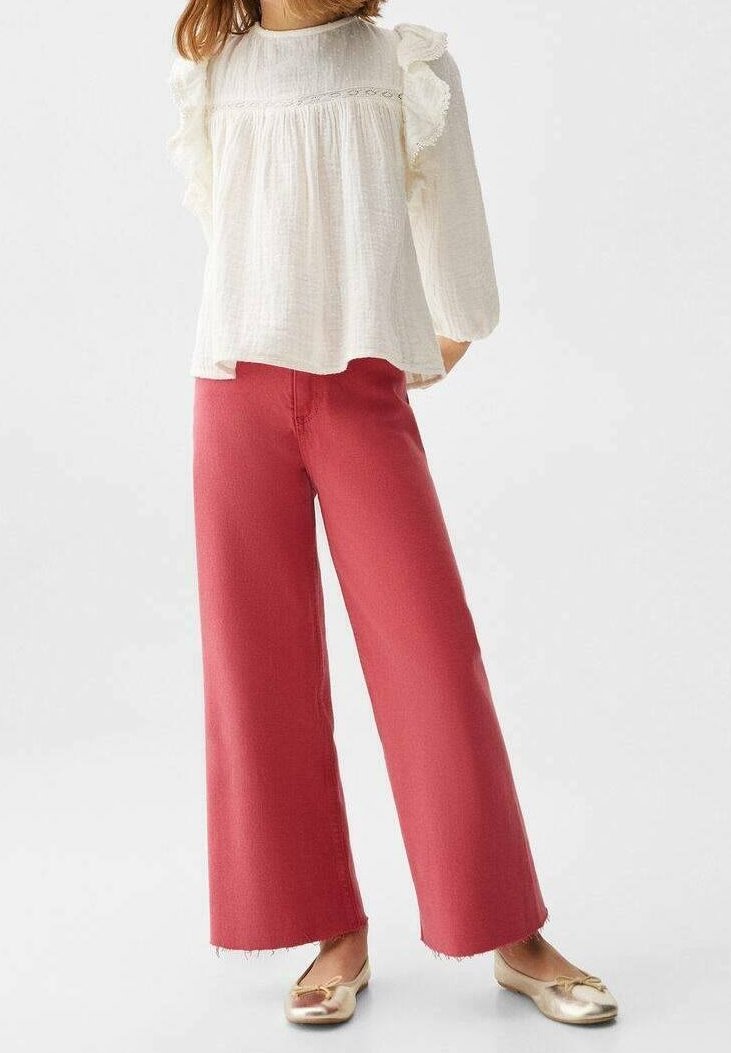 Ljusgräddfärgad blus med volanger och långa ärmar, kombinerad med vida, rosa byxor. Modellen bär blanka, gyllene platta skor. En enkel, avslappnad outfit.