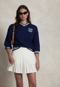 Hanorac bleumarin cu dungi albe și logo brodat, combinat cu o fustă plisată albă. Ochelarii de soare și o geantă de umăr completează look-ul.
