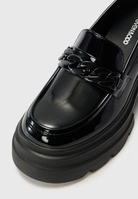 Mocassins en cuir verni noir avec une épaisse semelle en caoutchouc noir et un détail en chaîne sur l'empeigne. Texture lisse avec une finition brillante.