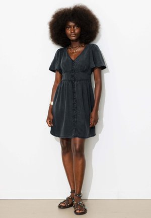 Femme debout contre un mur uni, portant une robe noire à manches courtes, longueur genou, des colliers superposés et des sandales à clous.