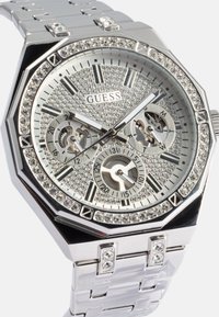 Relógio octogonal prateado com um bezel cravejado de cristais e um mostrador texturizado. As características incluem três submostradores e uma bracelete metálica.
