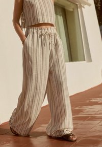 Personne portant un pantalon beige à jambes larges avec des rayures verticales et un crop top assorti, debout sur des carreaux de sol en terre cuite près d’un mur blanc.