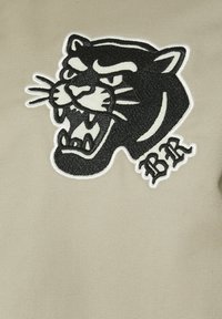Tête de panthère noire brodée avec dents découvertes et initiales "BR" sur fond de tissu beige.