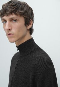Donkergrijze geribbelde trui met een hoge kraag en ritsdetail. Zachte textuur en aansluitend ontwerp benadrukken een moderne minimalistische stijl.