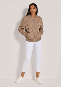 Vrouw in een beige bomberjack met handen in de zakken, witte cropped pants en witte sneakers, staand tegen een lichte achtergrond.