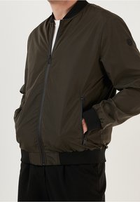 Tumman oliivin värinen pommikaukalotakki, jossa on sileä pinta, vetoketju, sivutaskut sekä ribatut hihansuut ja helma. Hihassa on logoyksityiskohta.