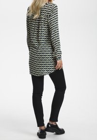 Tunique à manches longues avec un motif géométrique bleu marine, vert et crème. Coupe ample avec un ourlet arrondi. Associée à un jean slim noir et des chaussures.