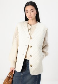 Mönstrad, krämfärgad väst med diagonala diamantmönster, med rund halsringning, knappar framtill och fickor, ovanpå en stickad, beige tröja.
